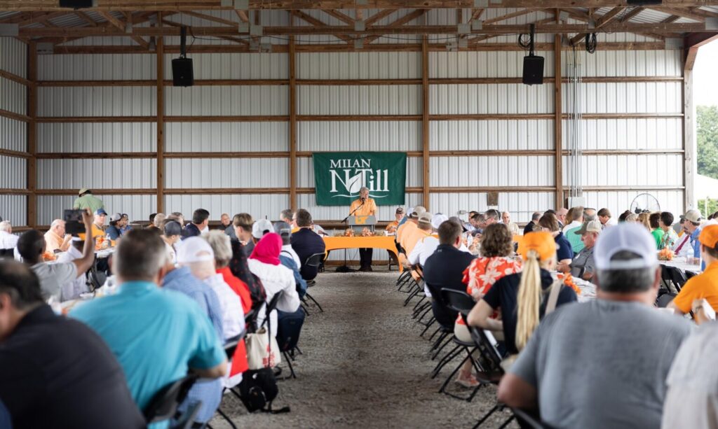 Tables of attendees listen to a speaker at a podium with a Milan No-Till banner