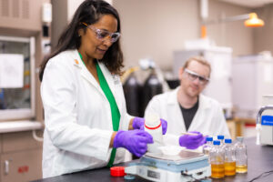 A scientist in a lab coat and safety goggles carefully measures a substance on a balance scale while another scientist works in the background.